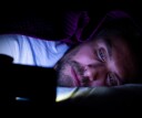 Bearded young man is lying in bed under his blanket. He cannot sleep and is watching something on his mobile phone.
