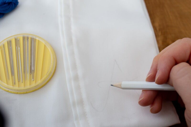 A hand holds a white pencil over a standard white pillowcase, where the person has lightly sketched a letter A into the fabric. Use this guide to learn how to embroider an initial into a pillowcase.