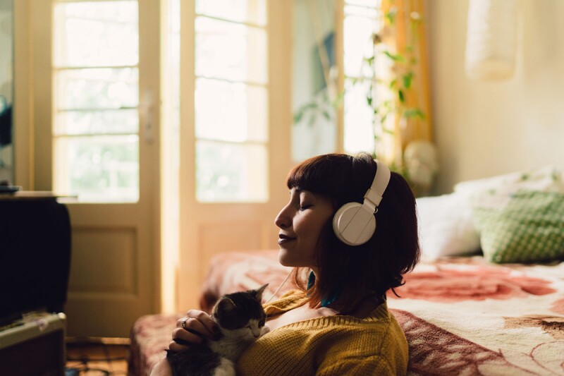 A woman sits on the floor in her bedroom with her eyes closed and a large set of headphones covering her ears. she is holding a cat. The bedroom fits perfectly with 2022 interior design trends. 