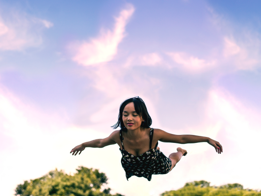 A girl flying in the air. The sky is pink and purple behind her and green trees below her.