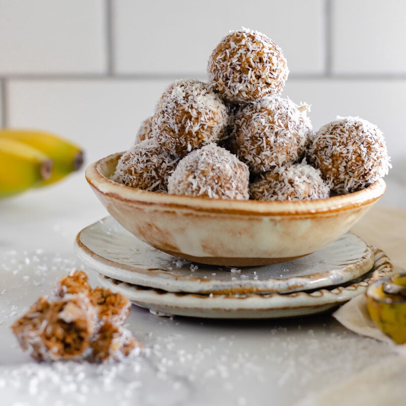A bowl of almond butter banana bites sits on a saucer with dustings of shredded coconut all around and whole bananas peeking into the frame in the background. Use this recipe as part of a sleep diet.