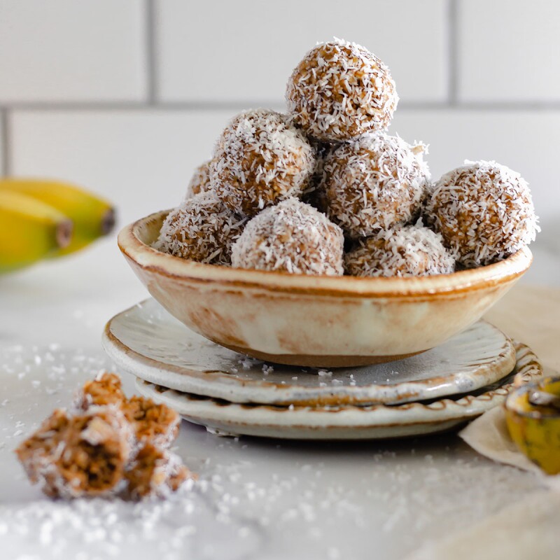 A bowl of almond butter banana bites sits on a saucer with dustings of shredded coconut all around and whole bananas peeking into the frame in the background. Use this recipe as part of a sleep diet. 