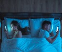 A couple doomscrolling in bed while basked in blue light from their cell phones.