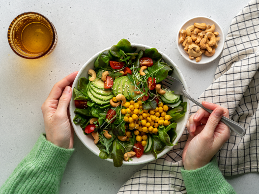 Anonymous female dining healthy vegan plant-based salad in bowl with avocado, cashew, micro-greens, pok choi, chickpeas, tomato, lettuce, cucumber, sesame. Flexitarian fatty acids and dietary fiber.