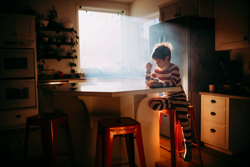 A young boy sitting a kitchen table eating cereal while the early morning light shines through the window.