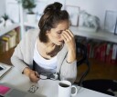 A young woman sits at her home at desk, holding her glasses with one hand and gripping her forehead with the other. She is suffering from a headache due to burnout syndrome, which also impacts sleep.