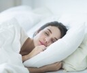 A woman sleeps peacefully in a bed of white linens with the perfect pillow supporting her neck. Getting a pillow fitting can help reduce pain and get a good night's sleep.