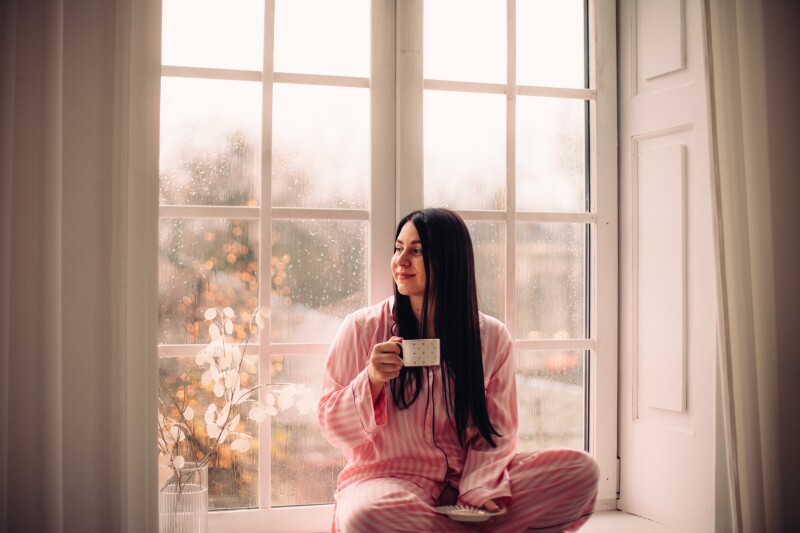 Young woman drinking coffee while sitting on the window sill at home during rainy weather