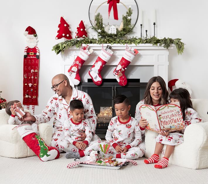 A family of four kids and two parents enjoy a holiday morning in matching family Christmas pajamas. The mother and daughter are reading a book, the father is interacting with the youngest son, and the two other sons are focused on their gifts.