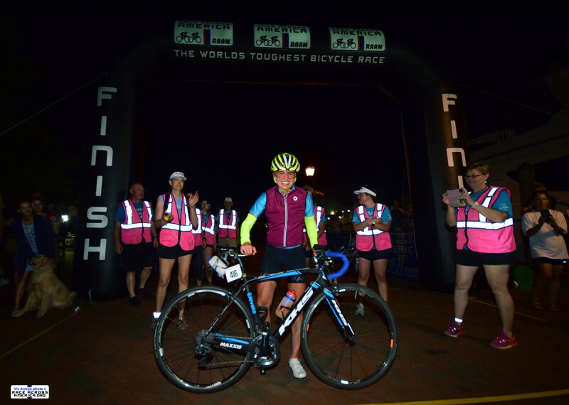 A winning cyclist shows off her post-race glow after completing Race Across America, or RAAM. She is surrounded by volunteers in pink safety vests clapping for her. She is looking forward to a great night of sleep!