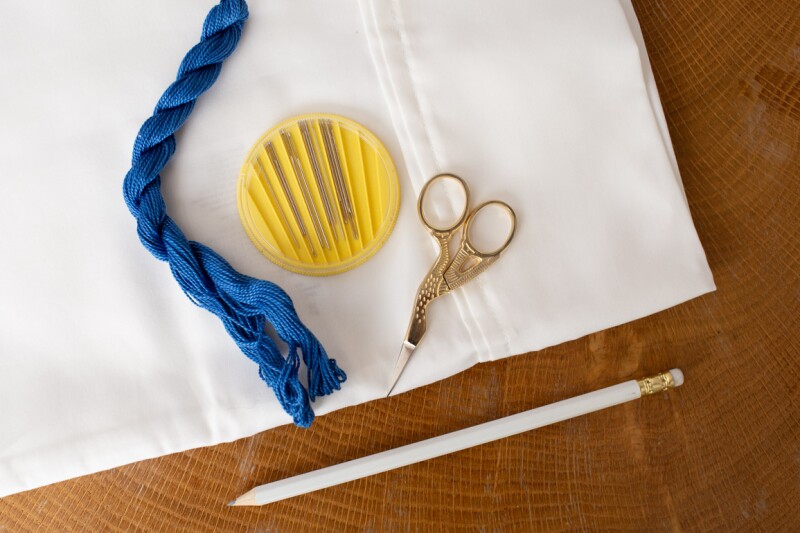 A set of embroidery supplies sits on a table: a standard white pillowcase, a pencil, scissors, needles, and fabric. Use this how to guide to embroider a pillow for unique, hotel-like home decor.