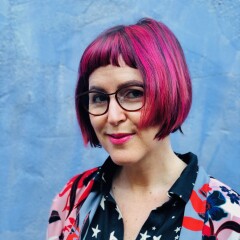 Headshot of writer and editor Jess Barron. She has short pink hair and glasses and is standing in front of a blue wall.