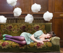 A young woman sleeping on a couch with dream clouds hovering above her.