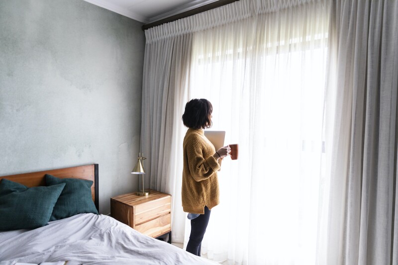 Person in cozy sweater holding a laptop and coffee mug while looking outside the window of their bedroom