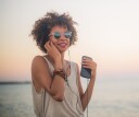 Person wearing headphones, sunglasses and bracelets, holding a phone and smiling in front of the ocean.